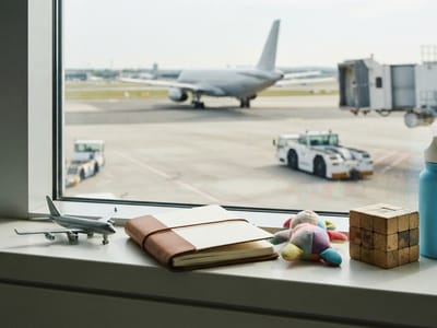 A windowsill holding a toy airplane, a leather-bound notebook, a plush toy, and a wooden puzzle cube, looking out an airport window at a plane and ground equipment on the tarmac.