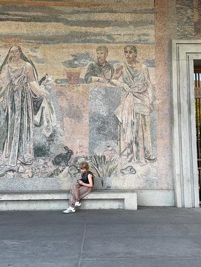A child sitting on a ledge beneath a large classical mosaic mural