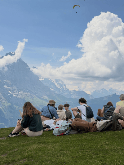 Group relaxing on a hilltop meadow with a paraglider above the Alps