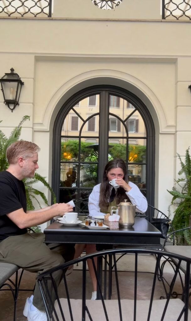 Two people enjoying coffee at an outdoor cafe table in front of an arched window, with ferns and classic Italian architecture in the background