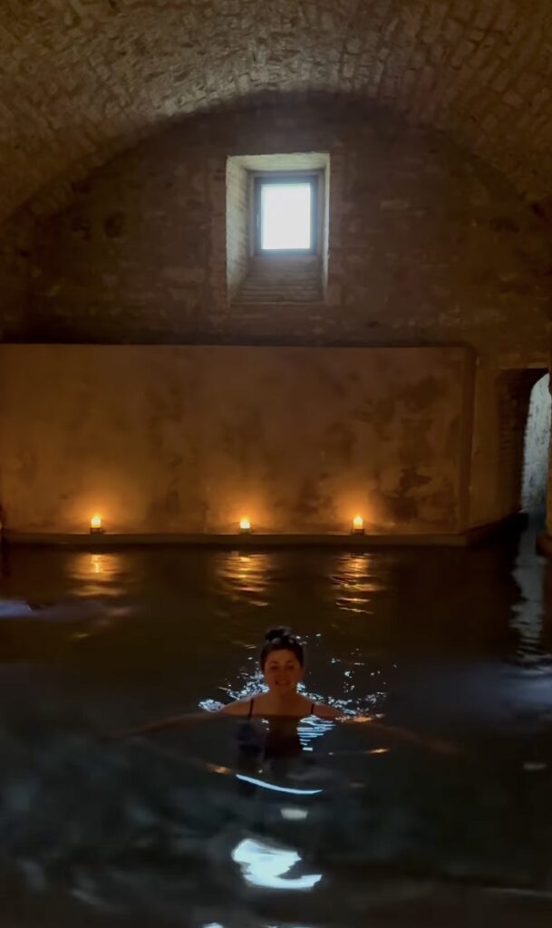 A woman soaking in a candlelit indoor thermal bath with stone vaulted ceiling and small arched window, Italy