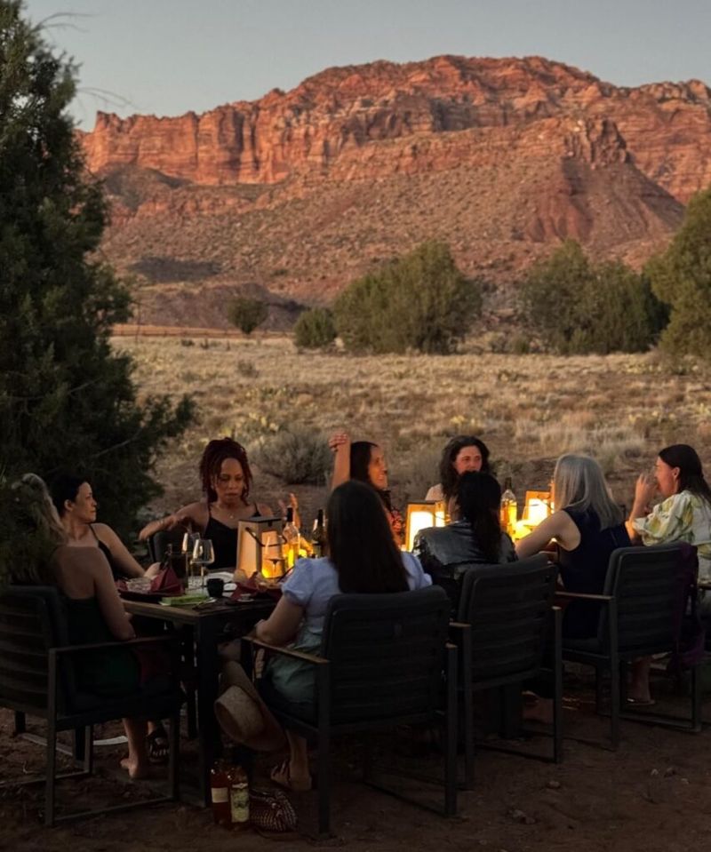 Group of people gathered around a candlelit outdoor dinner table at dusk, red rock canyon cliffs in the background, near Zion National Park