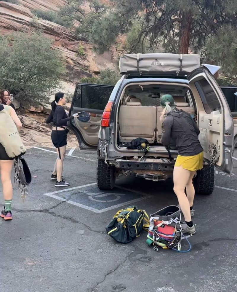 Three people unloading canyoneering gear from an SUV in a Zion National Park parking lot, red sandstone cliffs in the background