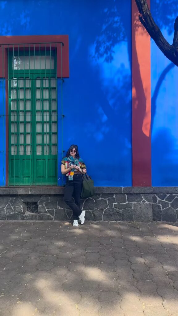 Jordan Ferney leaning against the cobalt blue exterior wall of Casa Azul, the Frida Kahlo Museum in Mexico City, with a green barred door and red trim behind her