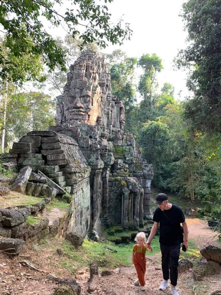 A man and young child walking hand in hand through a carved stone temple gate at the Angkor complex in Cambodia, with large stone faces carved into the tower above them
